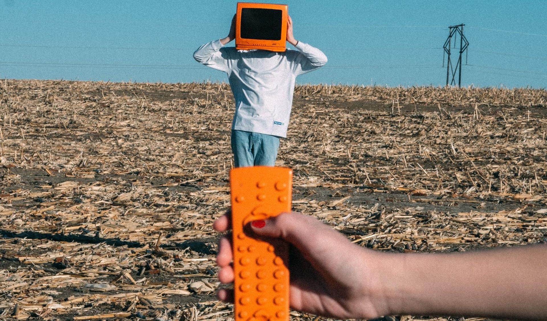 A person is standing on a harvested crops field and wears a TV on his head. A hand holds an orange remote control